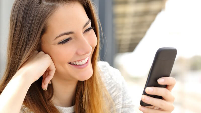 Junge Frau mit langen braunen Haaren nutzt lachend ihr schwarzes Smartphone