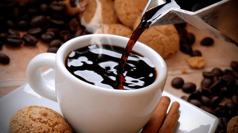 Frischer, dampfender Kaffee wird aus einer silbernen Kanne in die weiße Tasse gegossen, drumherum liegen Kekse