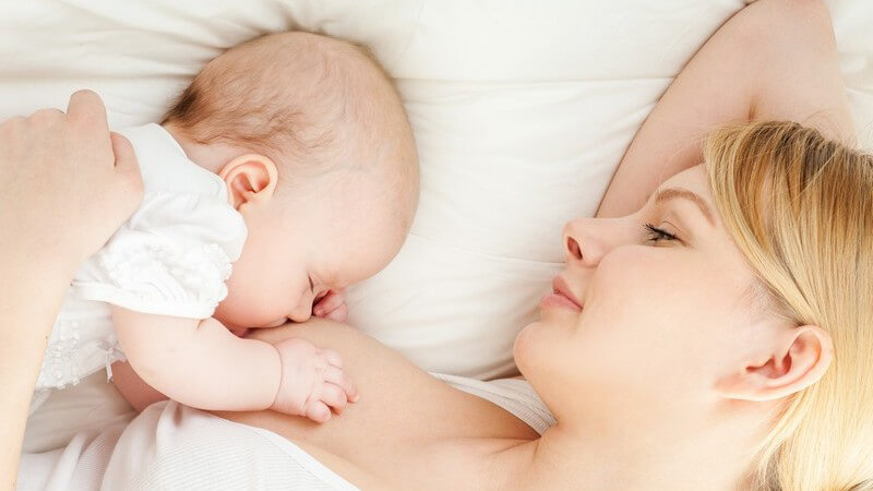 Junge Mutter mit blonden Haaren stillt ihr Baby im Liegen auf weißem Bett