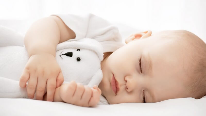 Baby liegt friedlich schlafend im Bett und hält ein weißes Kuscheltier in den Armen