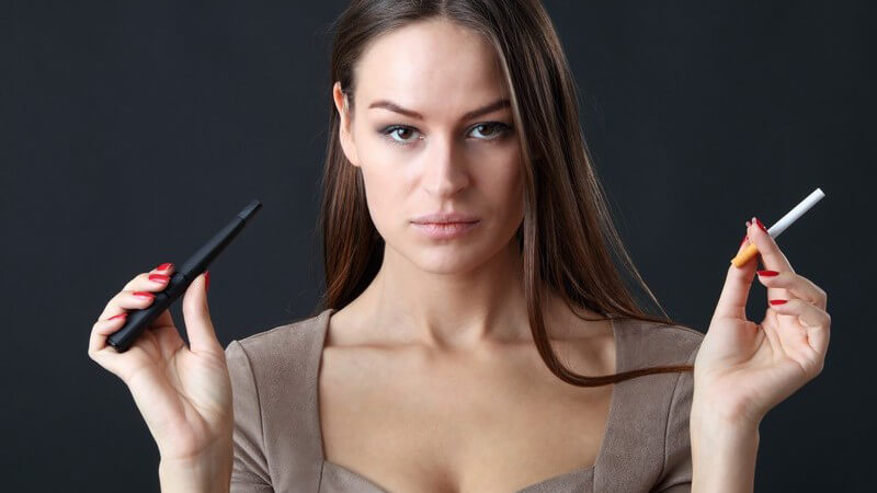 Dunkelhaarige Frau vor schwarzem Hintergrund hält in einer Hand eine E-Zigarette und in der anderen eine Tabakzigarette