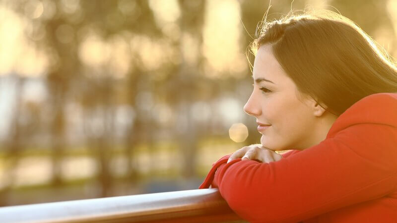 Junge Frau in roter Jacke lehnt bei Sonnenuntergang auf einem Geländer und blickt nachdenklich ins Leere