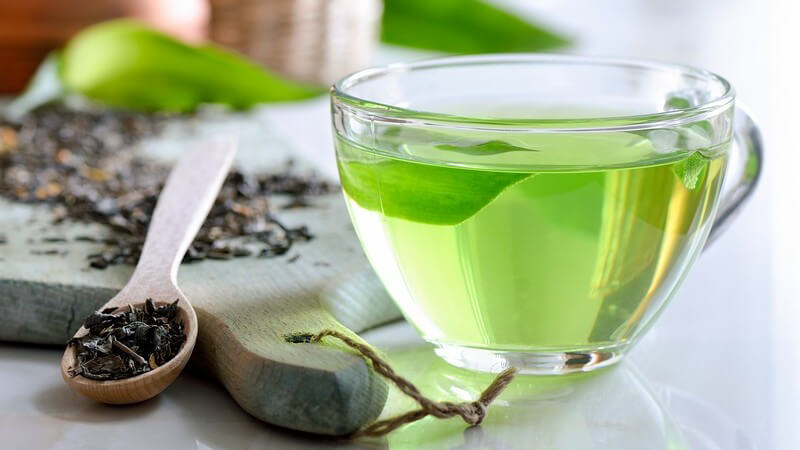Glastasse mit grünem Tee neben Holzbrett und -löffel mit getrocknetem Tee