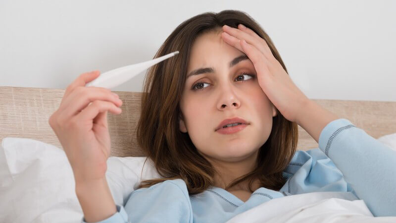 Braunhaarige Frau in hellblauem Pyjama liegt im Bett, fasst sich an die Stirn und blickt auf ein Fieberthermometer
