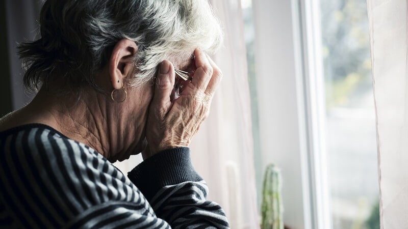 Seniorin mit Brille steht an einem Fenster und wirkt depressiv, die Hände vors Gesicht geschlagen
