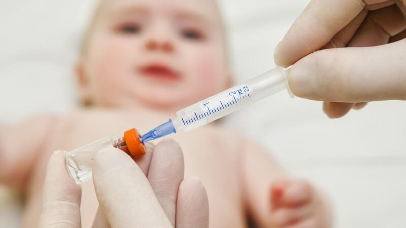 Arzt zieht eine Spritze aus einer Ampulle mit orangenem Deckel auf, im Hintergrund liegt ein Baby