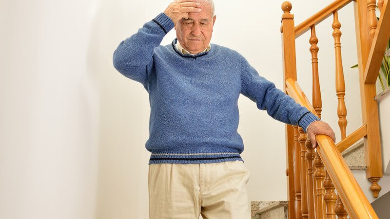 Alter Mann in blauem Pullover steigt eine Treppe hinab und fasst sich dabei an den Kopf wegen Schmerzen oder Schwindel