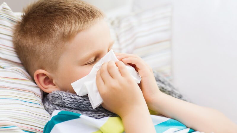 Junge mit Erkältung oder Grippe liegt mit Schal im Bett und putzt sich die Nase mit einem Taschentuch