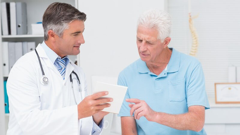 Arzt in weiß erklärt einem älteren Patienten in blauem Poloshirt ein Rezept