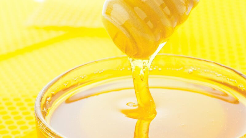 Honig fließt von einem Honiglöffel in ein fast volles Glasgefäß, gelber Waben-Hintergrund