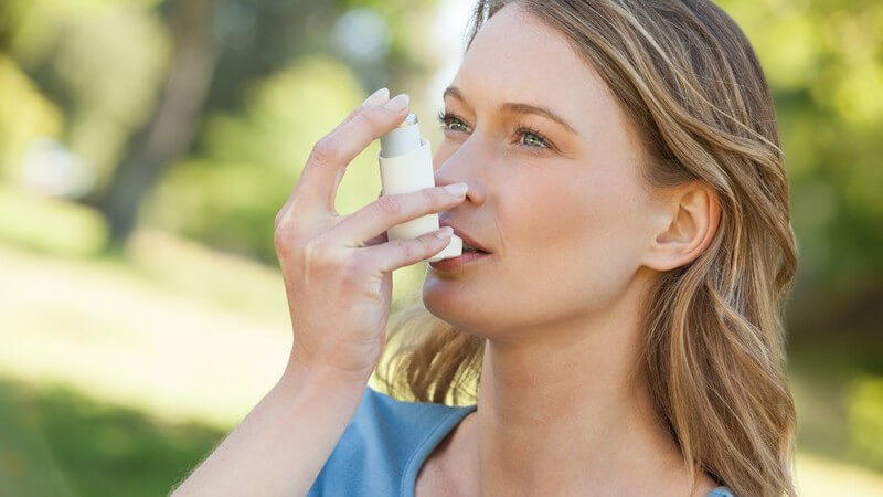 Junge Frau in blauem Shirt steht im Park und inhaliert aus einem Asthma-Inhalator