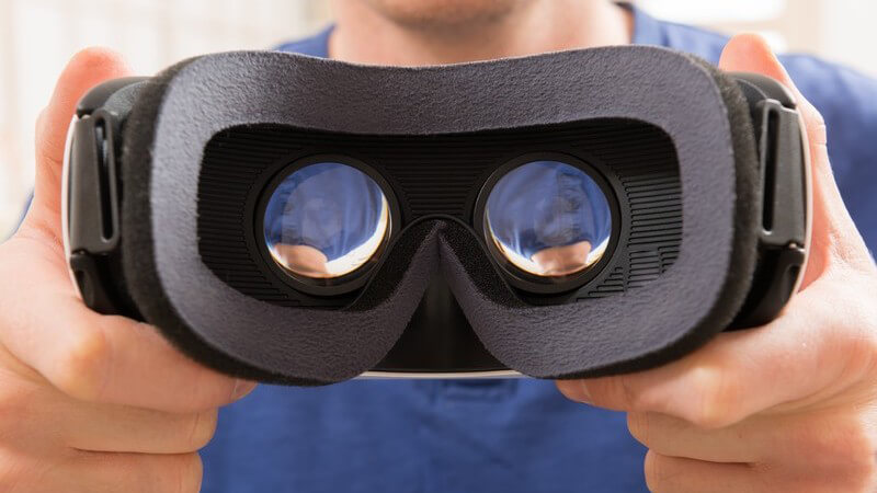 Mann in blauem Shirt hält eine schwarze Virtual Reality-Brille in die Kamera