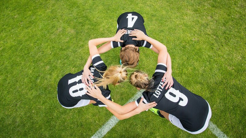 Drei Frauenfußballerinnen in schwarzen Trikots stehen auf dem Rasenplatz zur Besprechung zusammen