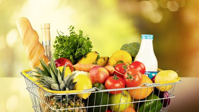 Einkaufswagen voll mit Lebensmitteln wie Obst, Gemüse, Milch und Baguettebrot, funkelnder Hintergrund