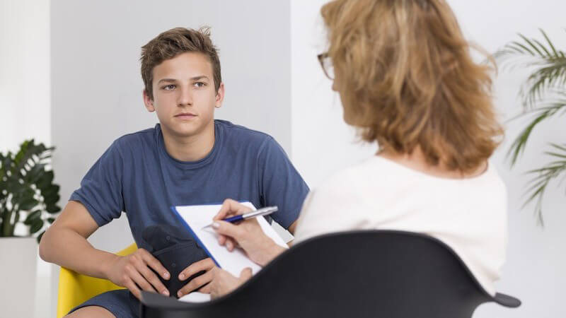 Junge in blauem Shirt sitzt in einer Sitzung bei einer Psychotherapeutin mit Brille, sie macht sich Notizen