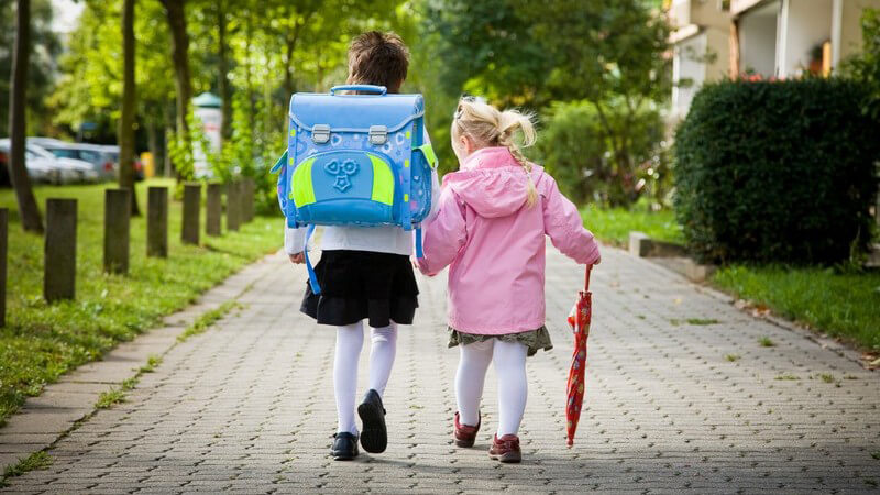 Rückenansicht Mädchen mit Schultasche an ihrem ersten Schultag mit kleinerer Schwester an der Hand
