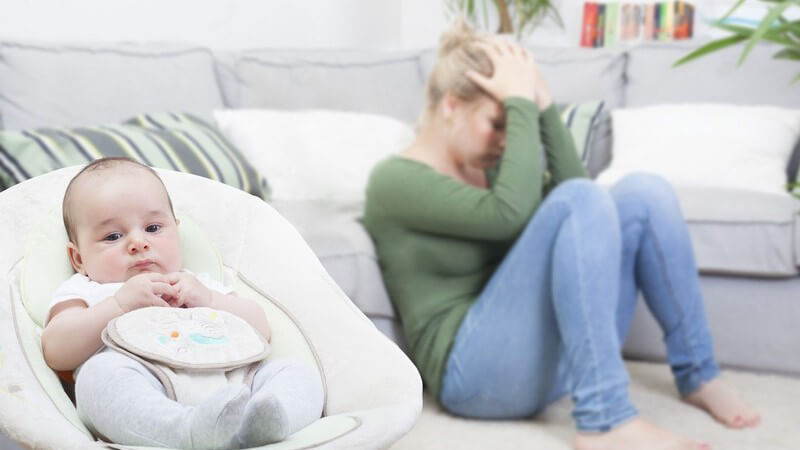 Depressive junge Mutter sitzt vor der Couch auf dem Boden, im Vordergrund sitzt ein Baby