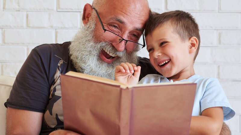 Großvater mit langem Bart und Brille liest seinem Enkelsohn ein Buch vor, beide lachen