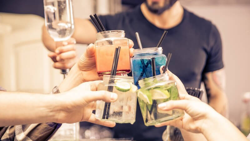 Personen stoßen mit breiten Cocktailgläsern mit schwarzen Strohhalmen an, im Hintergrund mixt jemand Cocktails