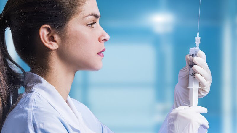 Junge Ärztin mit langen braunen Haaren und weißem Kittel bereitet eine Spritze vor, blauer Hintergrund