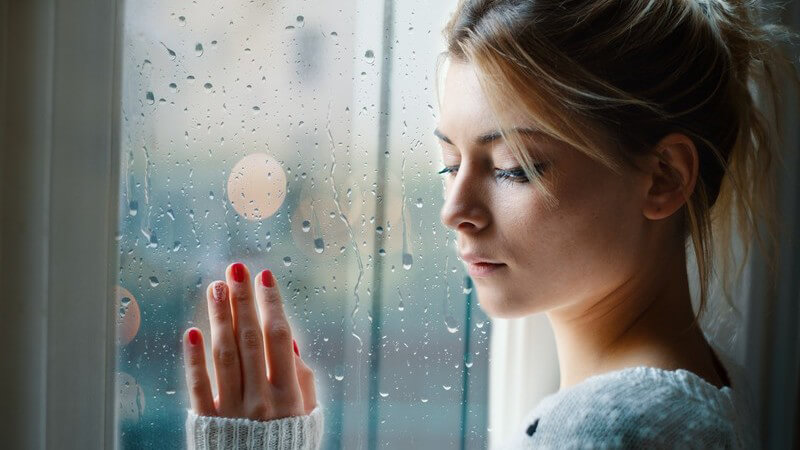 Junge Frau mit dunkelblonden Haaren und Wollpulli steht vor einer Fensterscheibe voller Regentropfen und guckt traurig