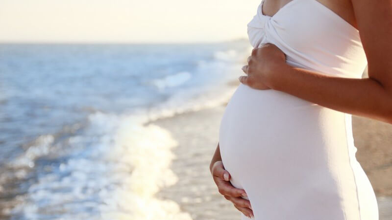 Schwangere Frau steht in weißem Sommerkleid am Strand und hält sich den Babybauch