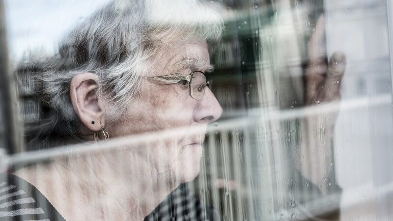 Seniorin guckt nachdenklich aus einem verregneten Fenster und wirkt depressiv