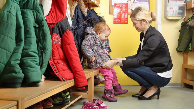 Blonde Mutter zieht ihrem Kind an der Garderobe im Kindergarten die Jacke an, um es abzuholen