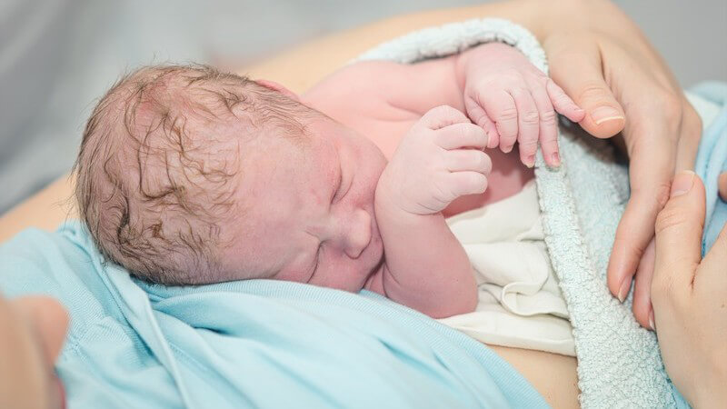 Neugeborenes Baby liegt kurz nach der Entbindung im Arm seiner Mutter in hellblauem Oberteil
