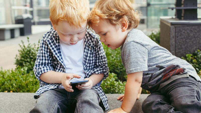 Zwei kleine Jungen sitzen draußen zusammen und befassen sich mit einem Smartphone