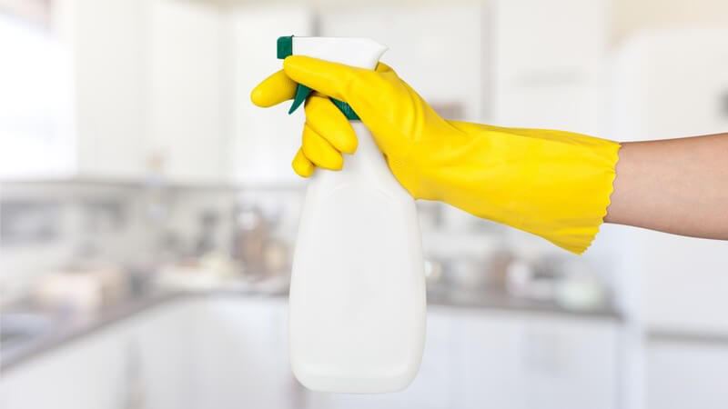 Hand mit gelbem Putzhandschuh hält weiße Sprühflasche mit Reinigungsmittel in die Luft, Küche im Hintergrund