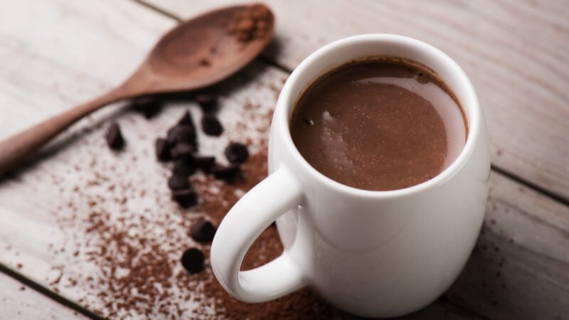 Tasse heiße Schokolade neben Kakaopulver und Holzlöffel auf Holztisch