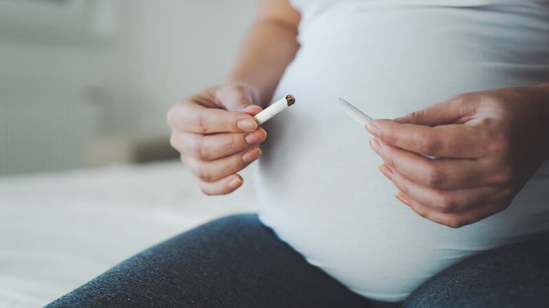 Schwangere in weißem Oberteil sitzt und hält eine zerbrochene Zigarette vor ihren Babybauch