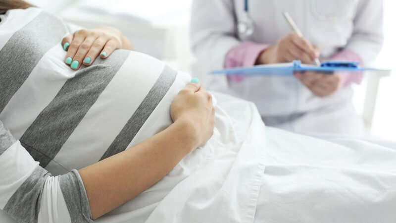 Schwangere Frau in grau-weiß gestreiftem Shirt liegt im Krankenhausbett und erhält Visite von einer Ärztin
