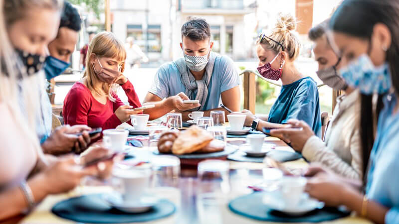 Junge Leute beim gemeinsamen Frühstück in einem Cafe, alle tragen Mundschutz und starren auf ihr Smartphone