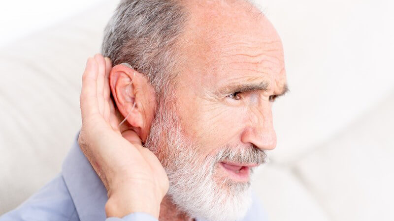 Alter Mann mit Vollbart trägt ein Hörgerät und hält die Hand hinters Ohr, um besser hören zu können