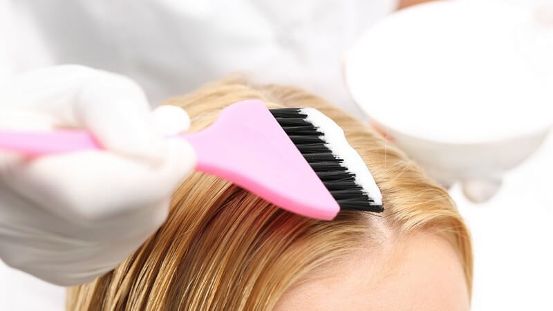Haarfärbemittel wird mit einem rosa Pinsel auf die bionden Haare einer Frau aufgetragen