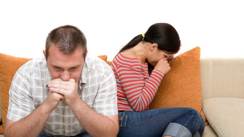 Junges Paar auf Couch, er sitzt vorne nachdenklich, sie von ihm abgewandt weint
