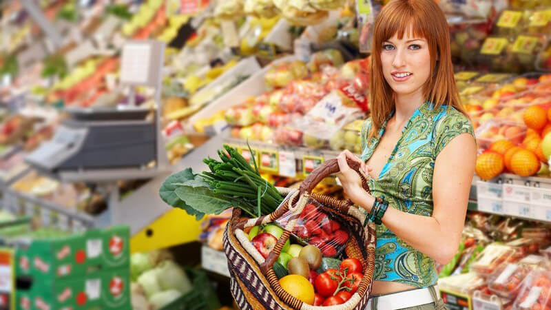Rothaarige Frau in grünem Outfit mit Obst-und-Gemüse-Korb in der Hand in einem Supermarkt