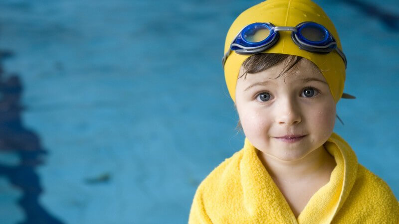 Kleines Mädchen mit Schwimmkappe, Handtuch und Taucherbrille vor Schwimmbecken