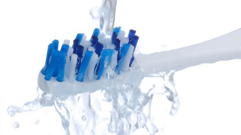 Nahaufnahme Kopf einer Zahnbürste mit weiß-blauen Borsten wird unter Wasserstrahl ausgespült, weißer Hintergrund