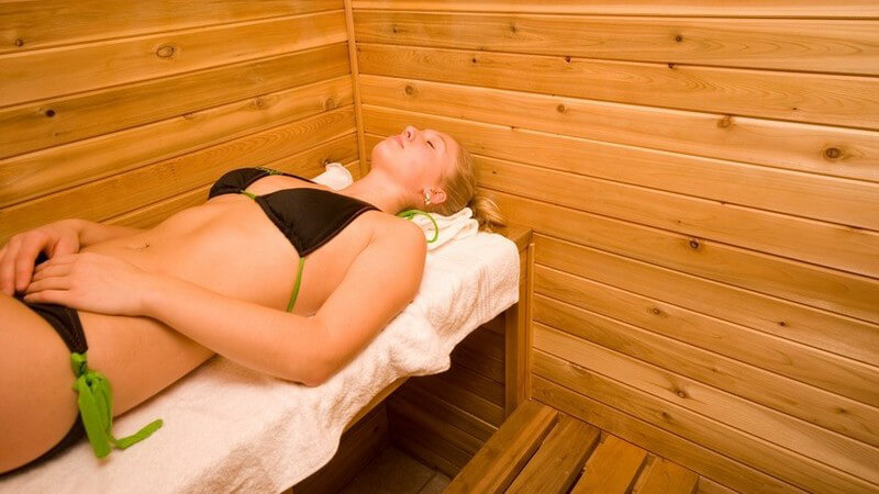 Blonde Frau im schwarz-grünen Bikini liegt auf einer Saunabank auf einem weißen Handtuch