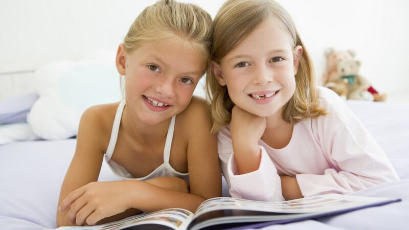 Zwei befreundete Mädchen im Schlafanzug liegen mit Buch zusammen im Bett, lächeln in Kamera