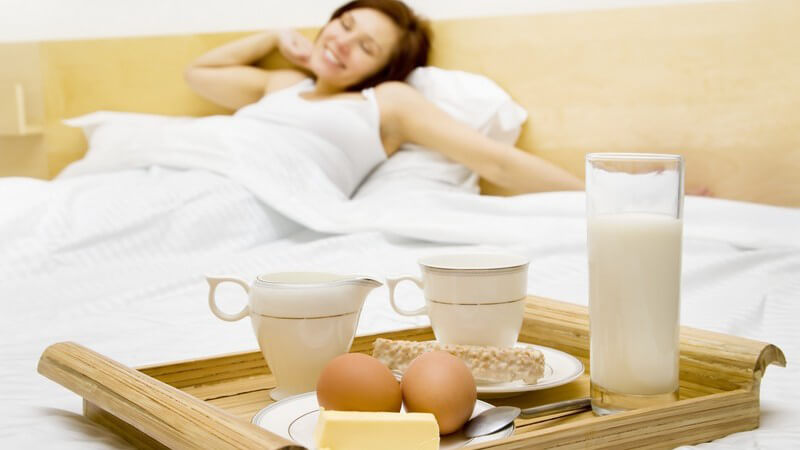Junge Frau streckt sich im Bett, vor ihr Tablett mit Frühstück