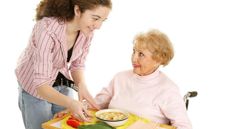 Junge Frau bringt alter im Rollstuhl sitzender Dame Tablett mit Essen