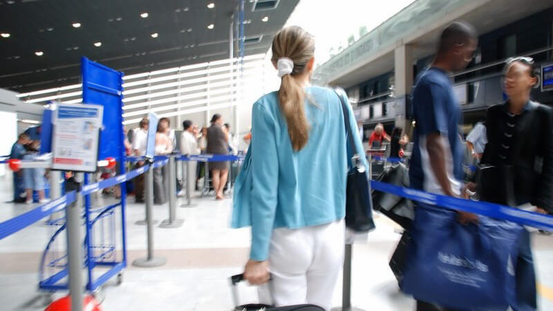 Rückansicht junge Frau zieht Koffer im Flughafen hinter ihr her, umgeben von vielen Menschen
