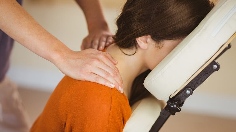 Frau in orangem Pulli sitzt bäuchlings auf einem Massagestuhl und wird an Nacken und Schulter massiert