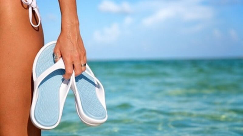Blau-weiße Flip-Flops vor hellblauem Wasser und Himmel, in der Hand gehalten von einer Frau neben Bein