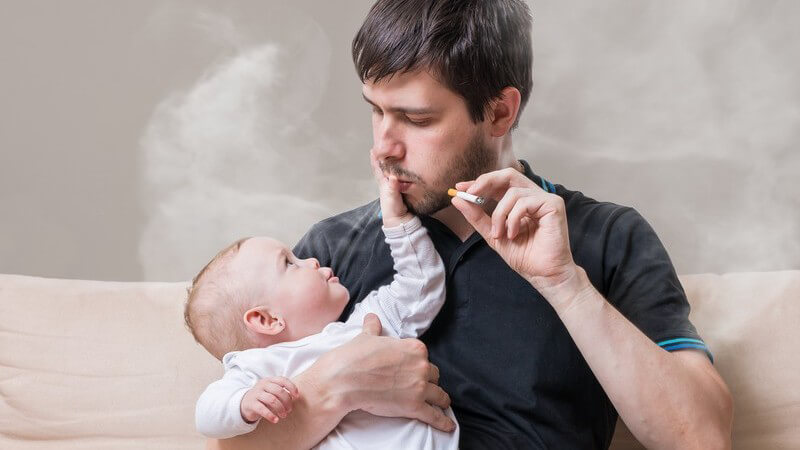 Baby auf dem Arm des Zigarette rauchenden Vaters hält ihm den Mund zu, umgeben von Qualm