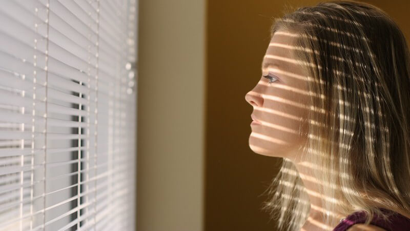 Junge Frau schaut bedrückt aus einem mit einem Plissee abgedunkelten Fenster